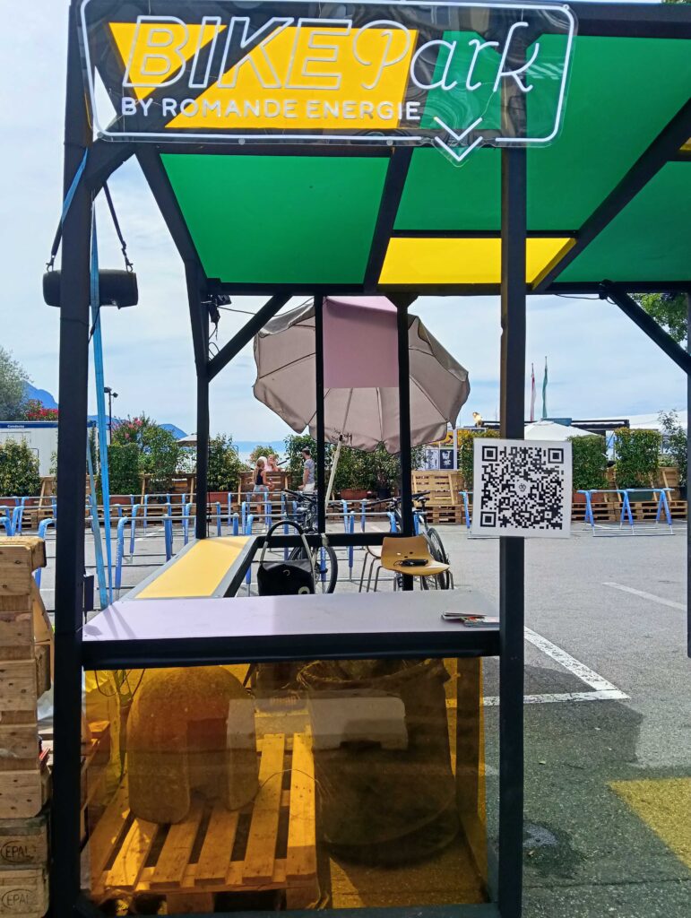 Kiosque de parking vélos en extérieur avec une enseigne néon « BIKEpark », des panneaux verts et jaunes, des palettes en bois et un panneau avec un code QR à proximité ; des personnes et des vélos sont visibles en arrière-plan.