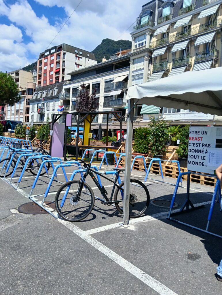 Rangée de vélos bleu vif garés dans un rack à vélos sur une place urbaine ensoleillée, avec des immeubles d’habitation et une colline en arrière-plan
