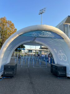 Outdoor event entrance under a white inflatable arch with 'Wheelskeep' branding, blue signage, and queue barriers.