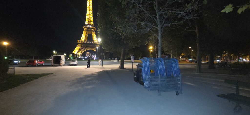 Vue nocturne de la Tour Eiffel illuminée en jaune, depuis un parc avec des chaises bleues empilées au premier plan.